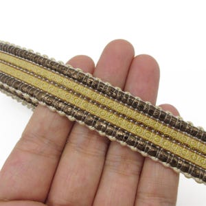 May include: A decorative trim ribbon featuring alternating horizontal bands of metallic bronze textured fabric and smooth gold-toned material with a repeating circular pattern, finished with small cream-coloured beaded edges along both sides.
