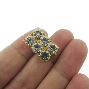 May include: A rectangular gold-coloured brooch with seven clear, faceted gemstones. The gemstones are arranged in a grid pattern, with a central row of three stones and two rows of two stones on either side. The brooch is set against a white background.