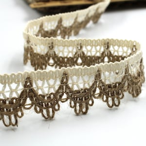 May include: A roll of decorative lace trim with a scalloped edge. The trim has a cream-coloured open weave top section and a brown, textured bottom edge. The lace is made of natural fibres and is suitable for various craft projects.