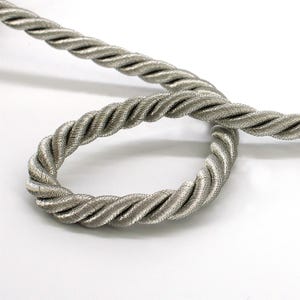 Puede incluir: Un cordón trenzado de color plata metálica con una estructura de cuerda de múltiples hilos y un brillo sutil. El cordón está enrollado para mostrar su textura en espiral uniforme y su grosor constante, destacando el acabado reflectante.