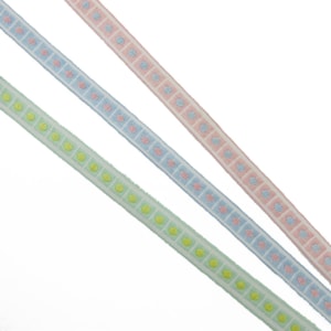 May include: Three decorative ribbons in pastel shades of green, blue, and pink. Each ribbon has a repeating square pattern with small, colourful designs. The ribbons are angled across the frame, highlighting their decorative use.
