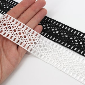 Puede incluir: Primer plano de un ribete de encaje blanco y negro. El encaje blanco presenta un patrón repetitivo de círculos y rectángulos. El encaje negro tiene un diseño similar. El encaje se sostiene en una mano, mostrando la textura y el detalle de la tela.