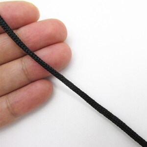 May include: A thin, black, braided cord with a tight, uniform weave pattern. The cord is round in cross-section and features a consistent, smooth texture throughout its length, showing a durable construction suitable for various crafting applications.