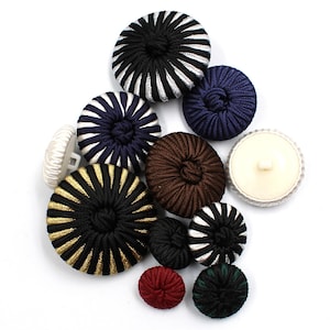 Può includere: Una collezione di bottoni decorativi in vari colori e design. I bottoni includono nero, bianco, blu navy, marrone, oro e rosso. Alcuni bottoni hanno un motivo a righe, mentre altri sono a tinta unita. I bottoni sono rotondi e hanno un aspetto strutturato.