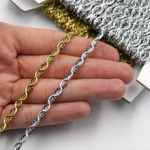 Może przedstawiać: Dwie szpulki metalicznej taśmy ozdobnej, jedna złota, a druga srebrna, z plecionym wzorem. Taśma jest pokazana w dłoni, z końcami taśmy wystającymi na zewnątrz.