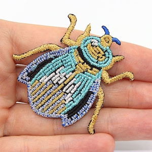 Może przedstawiać: Naszywka z haftowanym motywem stylizowanej pszczoły w kolorze niebieskim, złotym i czarnym.