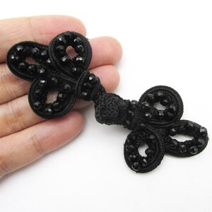 May include: Black frog closure with a decorative design. The closure features a series of loops and knots, with small, black, faceted beads. The closure is made of a woven material.