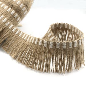 May include: Close-up of a roll of natural jute fringe trim. The trim has a woven edge with white rectangular accents and long, loose, brown fringe. The material appears to be a natural fibre, suitable for various craft and design projects.