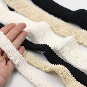 Puede incluir: Primer plano de varios adornos de flecos decorativos en varios colores. Los adornos incluyen blanco, negro y beige. El adorno blanco está sostenido por una mano. El fleco tiene una base texturizada y tejida con una fila de flecos.