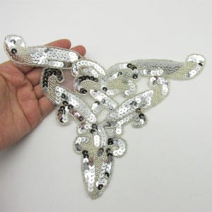 Puede incluir: Una aplicación decorativa con un patrón simétrico de remolinos, elaborada con lentejuelas plateadas y cuentas transparentes. El diseño es alargado con brazos curvos que se extienden hacia afuera, creando una forma compleja y reflectante.