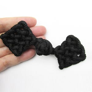 May include: A black Chinese-style frog closure fastener made of braided cord. The fastener features two diamond-shaped knotted ends connected by a central spherical knot, creating a decorative toggle mechanism for garments or textile projects.