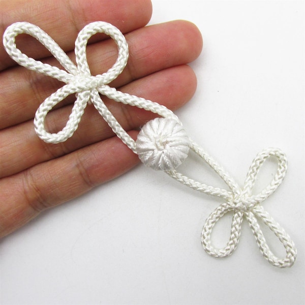 Puede incluir: Cierre de rana blanco decorativo hecho de cordón trenzado. El cierre presenta dos lazos en forma de flor y un nudo esférico central. Comúnmente utilizado en ropa y accesorios.