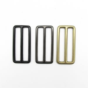 Puede incluir: Tres hebillas deslizantes rectangulares de metal dispuestas una al lado de la otra. La hebilla de la izquierda es de color negro mate, la central es de color gris plomo pulido y la de la derecha tiene un acabado de latón cepillado.