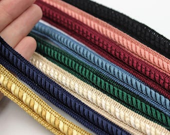 May include: A collection of decorative twisted cord trim strips arranged in a parallel stack. The colors include gold, navy blue, cream, emerald green, light blue, burgundy, peach, and black, each featuring a braided rope texture.
