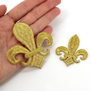 Pode incluir: Dois emblemas bordados de flor-de-lis na cor dourada com um brilho metálico. O emblema maior está posicionado sobre uma palma, enquanto o menor está colocado separadamente à direita. Ambos apresentam um trabalho de fio detalhado.