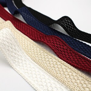 May include: Close-up of several woven ribbons in various colours. The ribbons include white, beige, burgundy, navy blue, and black. The ribbons have a textured, open weave design, suitable for crafts or embellishments.