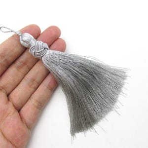 Può includere: Una nappa decorativa color argento caratterizzata da un denso ciuffo di fili sottili e dritti che pendono da una parte superiore in corda annodata. La nappa include un piccolo occhiello nella parte superiore per appenderla, con riflessi metallici.