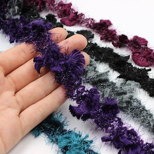 Puede incluir: Cinco carretes de ribete con una textura peluda y brillante. El ribete es en tonos de morado, negro, gris, azul verdoso y burdeos.