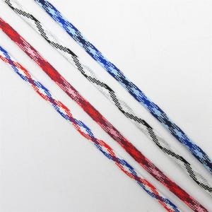Puede incluir: Tres cordones de diferentes colores con un patrón tejido. Los cordones son blanco con negro, blanco con azul y rojo con blanco y azul.