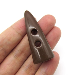 May include: A brown, horn-shaped button with two holes. The button is a solid colour and has a slightly glossy finish. It is held in a hand against a white background. This button is likely for sewing or crafts.