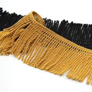 May include: Close-up of two decorative fringe trims. One is a golden yellow colour, and the other is black. Both trims have a twisted rope design with long, hanging fringe. The trims are likely used for embellishing clothing or home decor items.