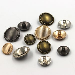 Könnte beinhalten: Eine Sammlung verschiedener Knöpfe im antiken Stil in Gold-, Bronze- und Silbertönen. Die Knöpfe sind rund und oval, mit strukturierten und glatten Oberflächen. Einige haben einen geriffelten Rand.
