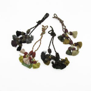 May include: Four decorative tassel ornaments featuring twisted cord loops and clusters of small, multi-coloured fabric tassels in shades of black, olive green, sage, beige, and deep burgundy, arranged in a fan-like formation on a plain white background.