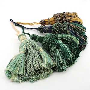 May include: A collection of four decorative tassels featuring intricate braided cord heads and long, multi-strand fringe skirts. The tassels are colored in various shades of green, blue, and gold, each attached to a matching braided hanging loop.