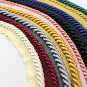 May include: Assortment of braided trim in various colours, including ivory, burgundy, grey, silver, gold, pink, and navy blue. The trim is made of a soft, woven material and has a rope-like texture.
