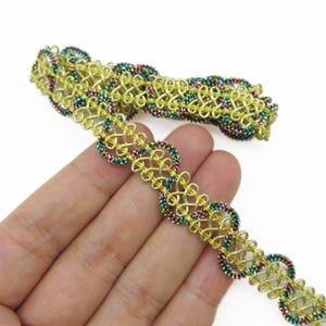 May include: Gold-coloured trim with an openwork design. The trim features a repeating pattern of interlocking shapes and is embellished with small, colourful beads in shades of teal, pink, and gold. The trim is shown against a white background.