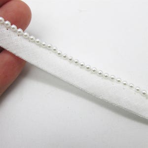 Peut inclure: Une bordure en tissu blanc ornée d'une rangée unique de petites perles artificielles blanches, rondes et brillantes, fixées le long du bord supérieur. La garniture présente une texture tissée et une finition droite pour les projets de couture.