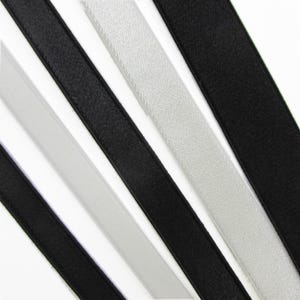 May include: A collection of smooth, narrow satin ribbons arranged diagonally. The set includes alternating strips of solid black and light silver-grey fabric, each featuring a consistent width and a subtle sheen across the surface of the material.