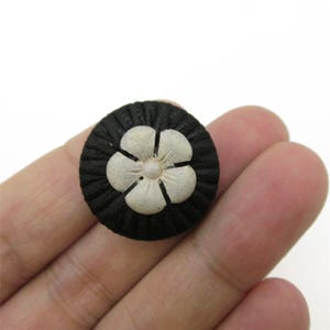 Pode incluir: Um botão redondo com uma base de tecido preto texturizado e plissado, apresentando um emblema floral de cinco pétalas de cor clara no centro. A flor tem um acabamento liso e mate, posicionado no meio do botão circular escuro.