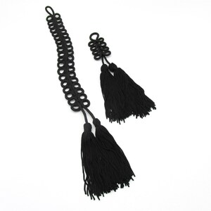 Puede incluir: Dos adornos decorativos de cordón negro con intrincados patrones de nudos y largas borlas de flecos fluidos. La pieza más larga tiene un diseño de bucles circulares repetitivos, mientras que la más corta presenta una estructura de nudos densa y multicapa.