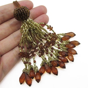 May include: A decorative tassel with a brown beaded top and strands of gold and brown beads. The tassel features teardrop-shaped amber-coloured beads at the bottom. The design is intricate and detailed, suitable for various craft projects.