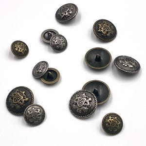 Puede incluir: Una colección de botones metálicos de estilo antiguo en varios tamaños. Los botones presentan un diseño de cresta heráldica en relieve con detalles intrincados. La paleta de colores incluye tonos de latón antiguo, plata y bronce, con un acabado texturizado.