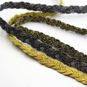 May include: Three braided cords in gold, black, and silver. The cords are woven together in a decorative pattern.