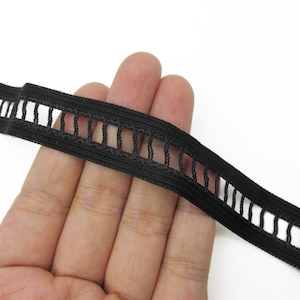 Puede incluir: Ribete elástico negro con una serie de recortes rectangulares a lo largo de un borde. El ribete se sostiene en una mano, mostrando la textura y el detalle del material tejido. El ribete probablemente se utiliza para proyectos de costura o manualidades.