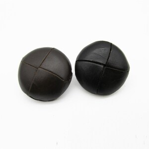 May include: Two dark brown, round buttons with a cross-stitch design. The buttons are made of a leather-like material and are set against a white background. These buttons are suitable for various sewing projects.