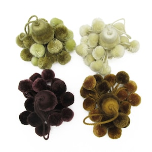 May include: Four decorative tassels arranged in a square formation. Each tassel features a central wrapped cord head with numerous small, soft pom-poms attached in shades of olive green, cream, deep burgundy, and warm golden brown.
