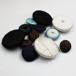Può includere: Una collezione di bottoni rivestiti in tessuto di varie dimensioni e colori. I bottoni includono nero, marrone, blu navy, azzurro e bianco, con una varietà di trame e design. Alcuni sono rotondi, altri ovali.
