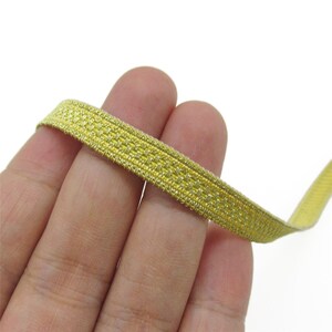 May include: A narrow strip of metallic gold elastic trim featuring a woven, textured pattern. The trim has a consistent width and a shimmering finish, displaying a repeating grid-like structure across its entire length and surface.