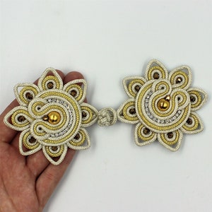 Könnte beinhalten: Zwei goldene und weiße Dekorverzierungen in Blumenform mit einem Perlenmittelpunkt. Die Verzierungen sind durch eine goldene Schnur verbunden.