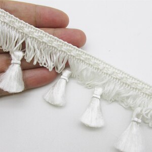 May include: A white decorative fringe trim featuring a woven header band with a repeating pattern, a row of short looped threads, and a series of evenly spaced, dangling white tassels attached along the bottom edge of the fabric trim.