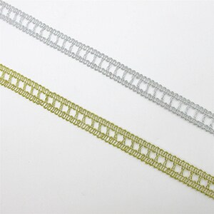May include: Two strips of decorative trim arranged diagonally across the frame. One strip is metallic silver and the other is metallic gold. Both feature a repeating openwork ladder pattern with thin, woven edges and a delicate, airy structure.