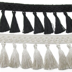May include: Close-up of two decorative trims. The top trim is black with a scalloped edge and black tassels. The bottom trim is silver with a scalloped edge and silver tassels. Both trims are made of a soft, silky material.