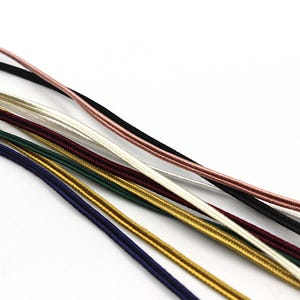 Può includere: Una collezione di sette cordini sottili e flessibili in vari colori, tra cui nero, oro rosa, bianco, bordeaux, verde scuro, oro e blu scuro. Ogni cordino presenta una consistenza liscia e fittamente intrecciata e un profilo cilindrico.