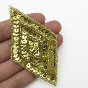 May include: A diamond-shaped applique patch featuring a dense arrangement of metallic gold sequins and small gold beads. The design is structured with concentric geometric lines of sequins and beads, creating a textured, reflective surface.
