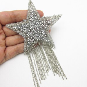 May include: A silver star-shaped embellishment with a beaded texture and dangling fringe. The star is heavily embellished with small, sparkling crystals, creating a glamorous and eye-catching design. The fringe is made of delicate silver chains.