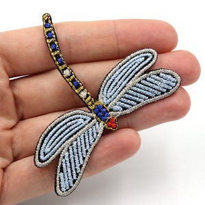May include: A blue and silver embroidered dragonfly applique with gold and red accents. The dragonfly has a long, thin tail and two pairs of wings.